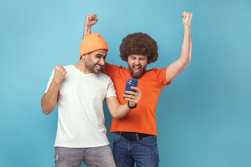 Portrait of two cheerful extremely happy young adult hipster men clenched fists while using mobile...