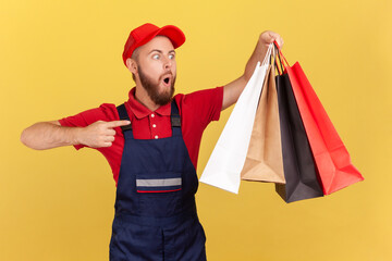 Portrait of surprised shocked delivery man in uniform pointing finger at shopping bags in his hand, fast delivery from mall, service industry. Indoor studio shot isolated on yellow background.