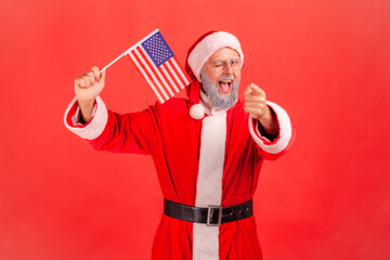 Satisfied elderly man with gray beard wearing santa claus costume waving USA flag winking and...