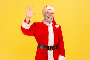 Positive good looking elderly man with gray beard wearing santa claus costume waving hand, greeting...