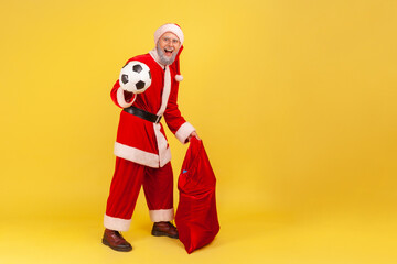 Full length of elderly man with gray beard wearing santa claus costume standing with soccer ball and big red bag with Christmas presents. Indoor studio shot isolated on yellow background.
