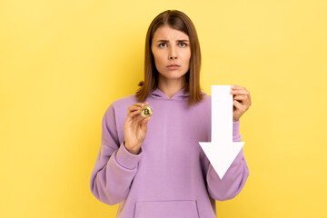 Portrait of unhappy upset woman showing bitcoin and white arrow pointing down, expressing negative...