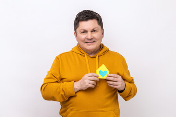 Long-awaited house purchase. Man holding small paper home with heart inside, satisfied with bought property, wearing urban style hoodie. Indoor studio shot isolated on white background.