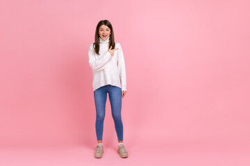 Full length portrait of excited female pointing finger aside, showing copy space for advertisement, wearing white casual style sweater. Indoor studio shot isolated on pink background.