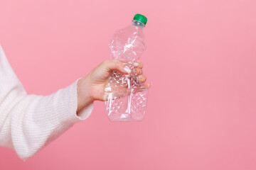 Female hand holding plastic bottle with green cap, sorting her rubbish, worrying about environment, wearing white casual style sweater. Indoor studio shot isolated on pink background.