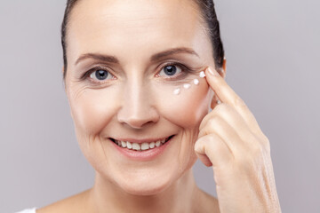 Closeup portrait of charming relaxed middle aged woman applies face cream, cares about complexion, touches cheek with hand, smiles gently at camera. Indoor studio shot isolated on gray background.