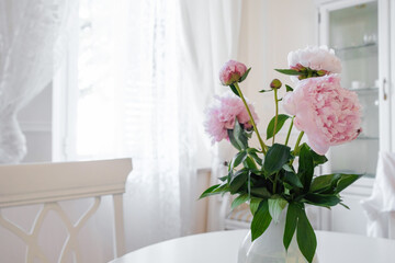 Bouquet of pink flowers in vase standing on table in room with white furniture. Peonies decorate apartment in spring at bright sunlight