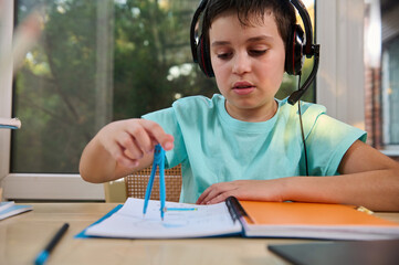 Portrait of an adorable schoolboy in audio headset, solving math problems using a compass to draw a circle in a notebook during an online geometry lesson. Back to school on new semester academic year