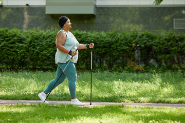 Side view full length of overweight black woman walking outdoors with nordic poles and enjoying cardio workout, copy space