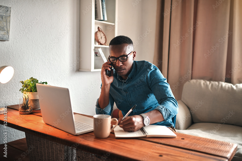 Poster Young entrepreneur on his phone while taking notes at his desk. Professional man working remote from home with technology. African American male has a business meeting on an audio call.