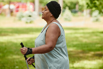 Side view portrait of overweight black woman walking outdoors with nordic poles and enjoying cardio workout