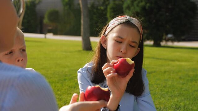 Schoolchildren of different ages are eating red apples on a green lawn. Close-up of a girl biting a red apple.