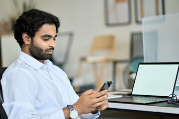 Obraz na płótnie Canvas Indian young business man company worker, professional employee, businessman executive holding smartphone using cellphone mobile corporate apps texting, checking financial data at work in office.