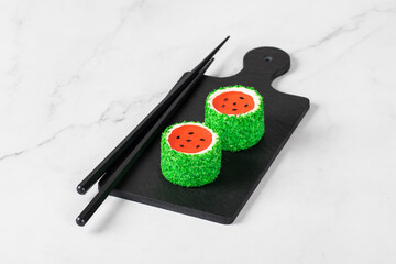 Two Children Mousse cream cakes with jelly watermelon in the form of round sweet Japanese sushi, sprinkled with green coconut chips, on a serving board. Light background