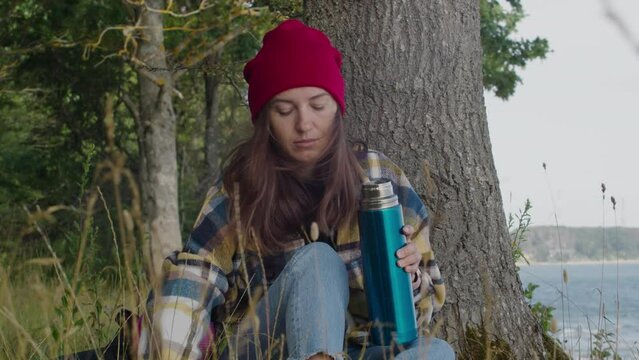 Woman hiker in hipster plaid shirt and red hat, backpacker traveler sit leaning against a tree and drinking hot tea from thermos and relaxing in forest. Happy travel wanderlust lifestyle