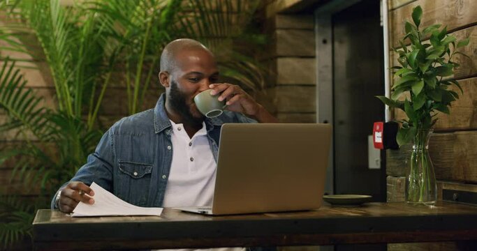 Multitasking entrepreneur checking paperwork and browsing on a laptop while drinking coffee in a cafe. Happy and cheerful male freelancer working remote analyzing and proofreading documents in a shop