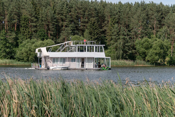 A ship in the form of a two-story houseboat floats along the river. Cruise on the river. Summer outdoor recreation.