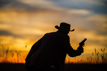 Silhouette of a cowboy in a hat with a revolver against a dramatic sunset sky. Western concept....