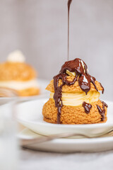 Craquelin Choux biscuits with cream vanilla filling pored with chocolate on light grey background. Traditional french pastry and dessert