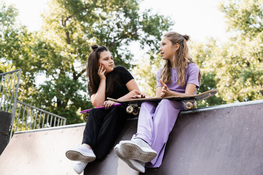 Sporty children girls with skateboard and penny boards are sitting and chatting with each other on sports ramp on playground. Friends communication and friendship. Sports extreme lifestyle.