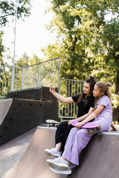 Children girls with skateboard and penny boards making selfie on phone on the sports playground. Children friendship concept. Kids friends smile and laugh and have fun together.