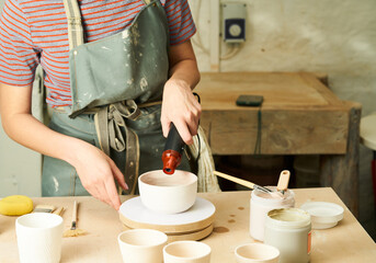 Close-up of girl painting clay mug with glaze. Woman coloring pottery in workshop with a paintbrush. Painter in green apron glazing clay pot.