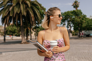 Fototapeta na wymiar Stylish young caucasian woman in sunglasses with coffee and magazines posing on street standing on exotic plants background. Successful adorable lady spend time outdoors