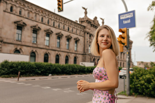 Modern pretty woman with short stylish hairstyle wearing summer dress is having fun in the city in warm sunny day. Attractive happy woman stands on background of old city building 