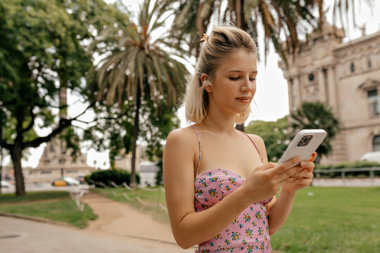 Stylish adorable woman with short blond hair in summer dress with floral print is scrolling smartphone stands on background of city park. Woman in wireless headphones is using smartphone 