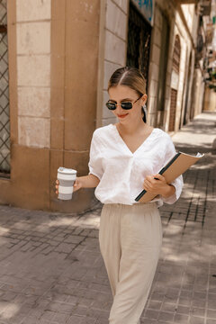 Outdoor portrait of attractive stylish pretty girl with blond hair wearing white shirt and beige pants in sunglasses walking on the street in sunlight with notes and coffee. Fashion, beauty