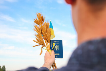 Defocus young man holding ukrainian passport, flag and wheat spikes of wheat tied and flag on the...