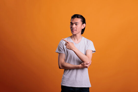 Young Smiling Confident Asian Man Pointing To Side With Forefinger, Advertising Product. Cheerful Teenager Standing, Showing Promotion, Studio Medium Shot On Orange Background