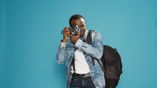 Cheerful photographer holding dslr camera to capture photo, taking professional picturs and carrying backpack. Photographing with lens on holiday vacation journey, standing in studio.