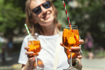 Portrait young adult happy smiling woman wear sunglasses enjoy park party holding in hand offer aperol spritz cocktail. Female person drink alcoholic beverage with friends resting lounge area outdoor