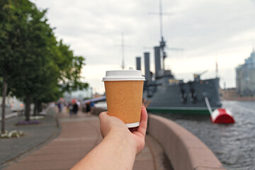 paper cup of natural coffee in women's hand on bridge of river next to ship cruiser Aurora in St....