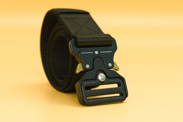 A black canvas belt with a black fast open metal male buckle and a black belt loop lays on the orange background. Security. Accessory. Fabric. Army. Tactical. Black. Metal. Iron. Fasten. Nylon. Yellow
