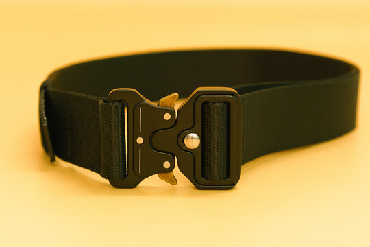 A front view of a black canvas belt with a black metal male buckle and a black belt loop lays on the orange background. Utility. Hiking. Material. Outfit. Safety. Soldier. Steel. Uniform. Weapon