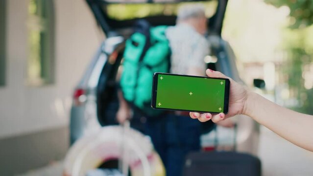 Person standing with touchscreen device having isolated template display while people loading luggage in car trunk. Woman standing with modern smartphone device having green screen mockup display.
