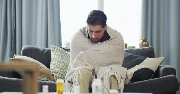 Sick, cold and tired young man with a fever sitting at home on sofa. Man showing symptoms of a flu, illness or virus in isolation shivering and feeling unwell having health problems.