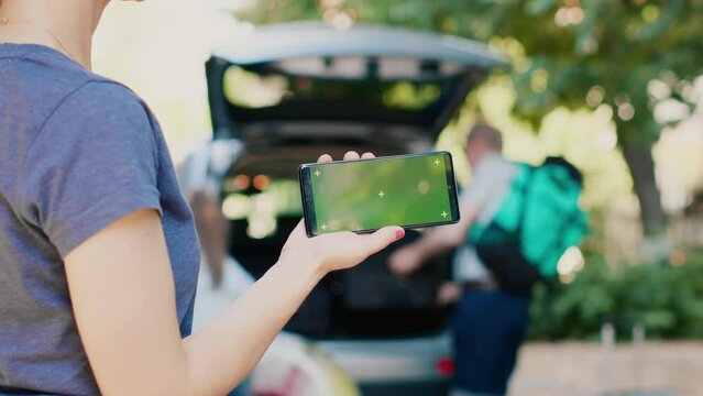 Person standing with phone having green screen mockup display while people in background getting ready for field trip. Woman standing with modern smartphone device having isolated template background.
