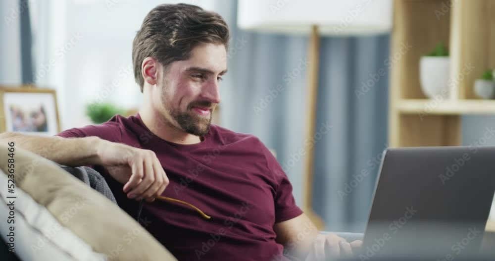 Poster satisfied man browsing on laptop and smiling after finishing work task while relaxing on the couch a