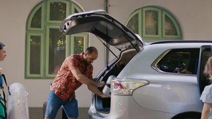 Casual family going on summer trip while loading luggage in vehicle trunk. Mother and father putting baggage and trolleys inside car while excited daughter getting ready for beach vacation.