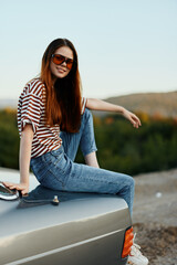 A fashion woman in stylish glasses, a striped t-shirt and jeans sits on the trunk of a car and looks at the beautiful nature of autumn