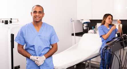 Successful experienced Hispanic cosmetologist in blue overall standing in modern aesthetic medicine office on background with female assistant checking equipments..