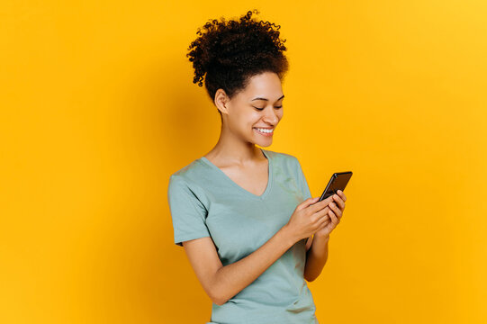 Satisfied glad african american young woman with curly hair, using her smartphone, looking at the screen, browsing the internet, chatting with friends, standing on isolated orange background, smiling