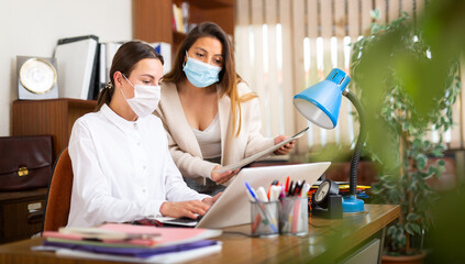 Woman assistant in face mask standing with documents near her female boss in office