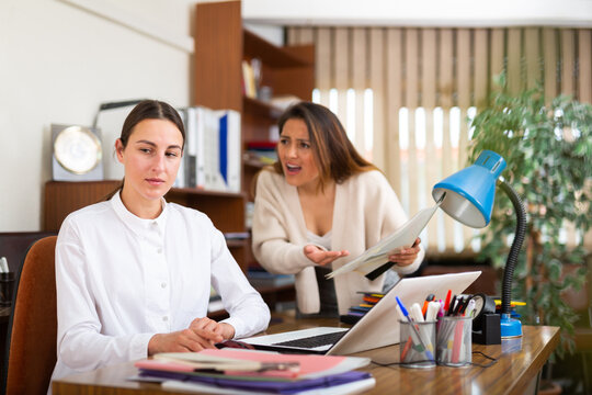Angry business woman criticizing sad frustrated female employee sitting at office desk at work