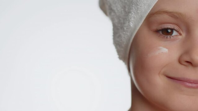 Young child girl applying cleansing moisturizing cream on cheekbones isolated on white background. Teenager kid face skin care healthy treatment, natural creme cosmetics. Perfect fresh clean skin