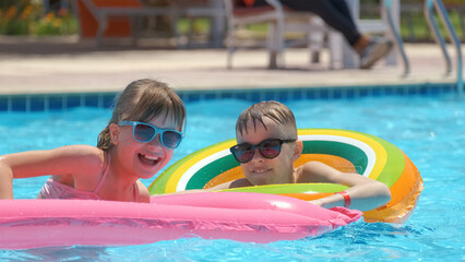Naklejka premium Happy children brother and sister playing on inflatable air mattress in swimming pool on sunny summer day during tropical vacations. Summertime activities concept