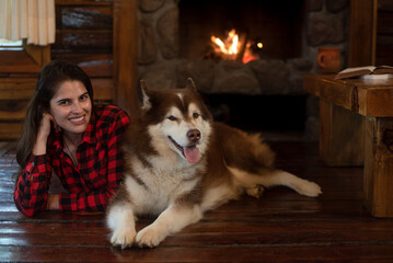 Woman with siberian husky dog sitting on the wooden floor in front of the burning fireplace...